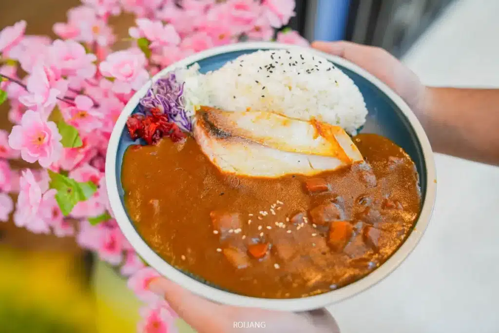 จานแกงกะหรี่ญี่ปุ่นเสิร์ฟพร้อมข้าว ปลาเผา และผัก จากร้าน JAPs ตะกั่วป่า วางอยู่หน้าดอกซากุระสีชมพู.