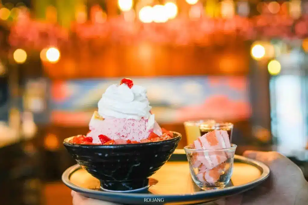 ชามน้ำแข็งไสราดด้วยวิปครีมและสตรอเบอร์รี่ เสิร์ฟพร้อมเค้กฟองน้ำขนาดเล็กสองถ้วยบนถาด ในร้านอาหาร JAPs ตะกั่วป่า ที่ตกแต่งด้วยแสงไฟอบอุ่น.