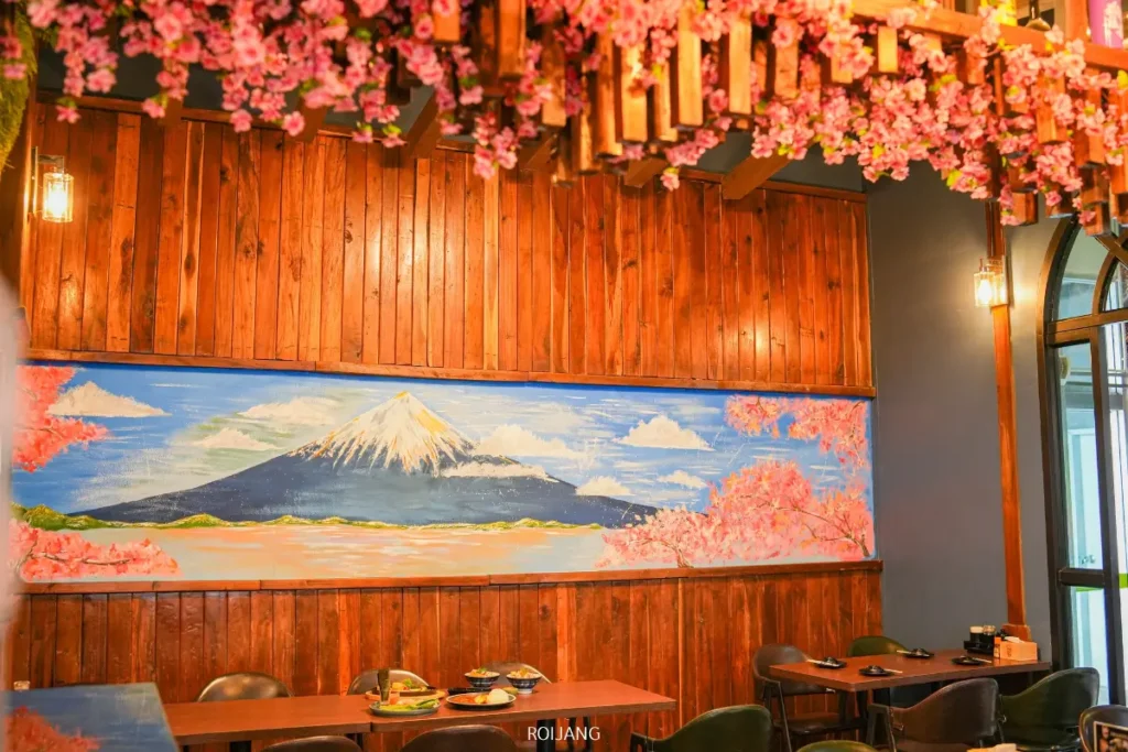 ร้านอาหารที่มีผนังไม้ ตกแต่งด้วยดอกซากุระ และภาพจิตรกรรมฝาผนังภูเขาฟูจิพร้อมต้นไม้สีชมพู JAPs ตะกั่วป่า มีโต๊ะและเก้าอี้ที่แสนสบายให้ลูกค้าได้นั่งเพลิดเพลินกับบรรยากาศที่สดใสนี้.