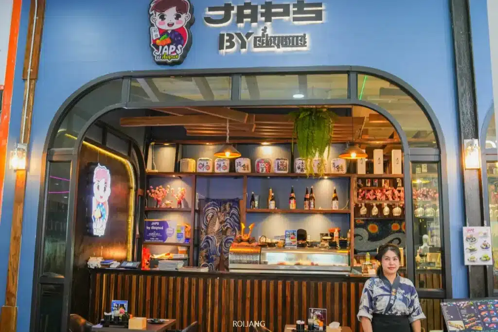 ร้านอาหารธีมญี่ปุ่น JAPs ตะกั่วป่า มีเคาน์เตอร์ ชั้นวางของตกแต่งและขวดต่างๆ ที่นั่งไม้ และพนักงานต้อนรับลูกค้าที่ทางเข้า.