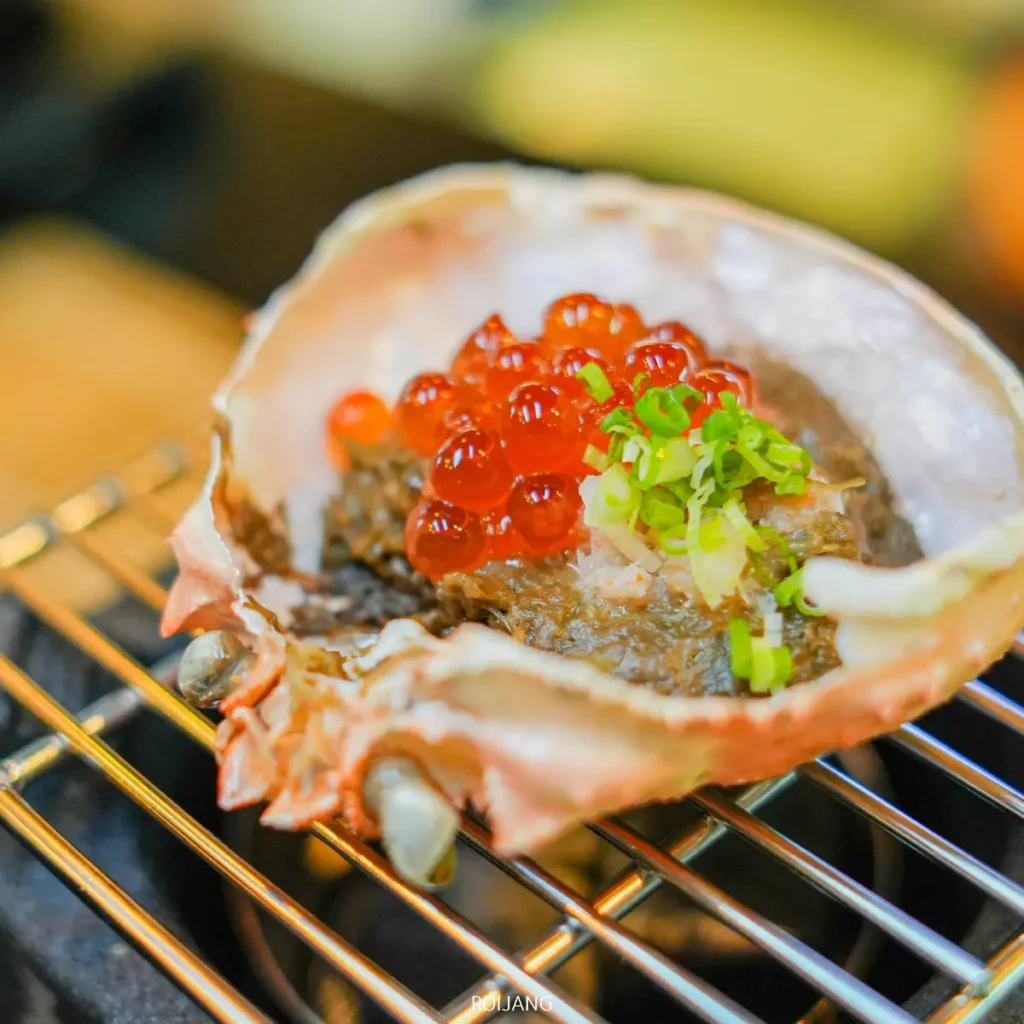 เปลือกปูที่เต็มไปด้วยเนื้อปู โรยด้วยไข่ปลาแซลมอนและต้นหอมซอย วางอยู่บนตะแกรงโลหะ—จานที่น่าลิ้มลองซึ่งได้รับแรงบันดาลใจจากรสชาติของ JAPs ตะกั่วป่า.