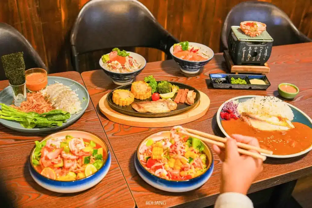 โต๊ะที่จัดวางอาหารญี่ปุ่นหลากหลายชนิดที่ร้าน JAPs ตะกั่วป่า รวมถึงสลัด ข้าวแกงกะหรี่ ปลาเผา ซาชิมิ และข้าวหน้าต่างๆ โดยมีมือกำลังถือตะเกียบอยู่ด้านหน้า.