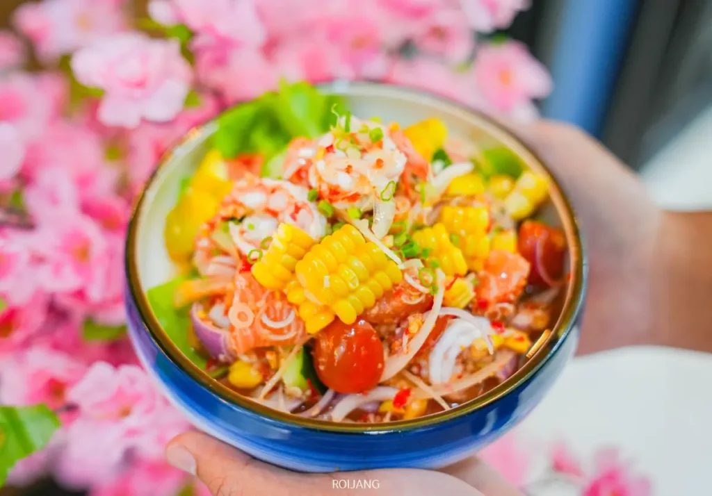 ชามสลัดไทยใส่ข้าวโพด มะเขือเทศเชอรี่ หัวหอม และผักสด ถือด้วยสองมือ โดยมีดอกไม้สีชมพูเป็นฉากหลังที่ JAPs ตะกั่วป่า.