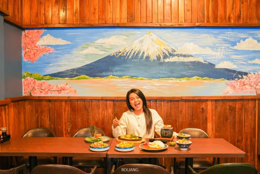 ผู้หญิงคนหนึ่งนั่งอยู่ที่โต๊ะพร้อมอาหารญี่ปุ่นหลากหลายชนิดที่ร้าน JAPs ตะกั่วป่า ยิ้มและทำสัญลักษณ์สันติภาพอยู่หน้าภาพจิตรกรรมฝาผนังภูเขาฟูจิภายในร้านอาหารที่อบอุ่นซึ่งมีผนังไม้.