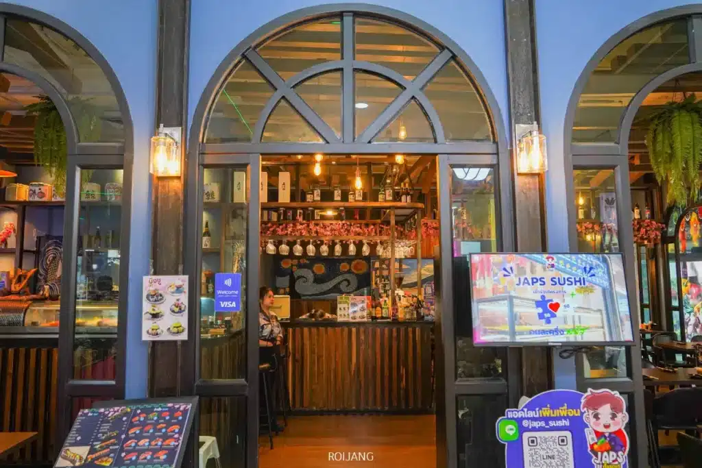 ภาพถนนหน้าทางเข้า JAPs ตะกั่วป่า ร้านซูชิญี่ปุ่น พร้อมเมนูที่จัดแสดง โคมไฟแขวน และของตกแต่งที่มองเห็นได้ภายในร้าน.