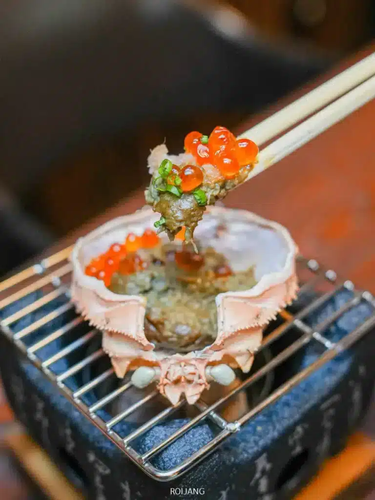 ตะเกียบคีบเนื้อปู ไข่ปู และเครื่องเคียงวางอยู่เหนือเปลือกปูบนเตาย่างขนาดเล็กที่ร้าน JAPs ตะกั่วป่า.