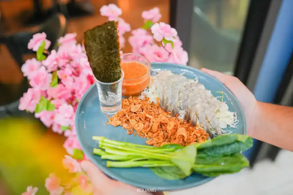 จานสีน้ำเงินจาก JAPs ตะกั่วป่า ประกอบด้วยปลาสดหั่นบาง, หอมทอดกรอบ, ผักใบเขียว, แผ่นสาหร่ายในแก้ว, และถ้วยเล็ก ๆ ของน้ำจิ้ม ทั้งหมดถูกถือด้วยสองมือ.