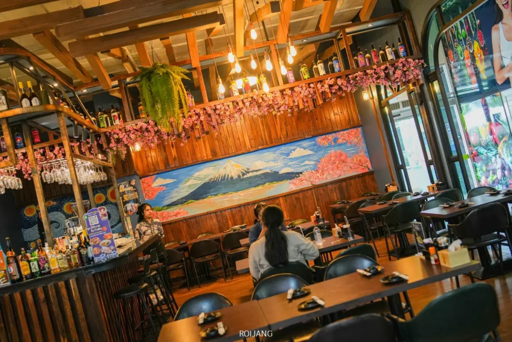 ผู้หญิงคนหนึ่งนั่งอยู่คนเดียวที่โต๊ะในร้าน JAPs ตะกั่วป่า ซึ่งเป็นร้านอาหารธีมญี่ปุ่น ตกแต่งด้วยไม้ โคมไฟแขวน และภาพจิตรกรรมฝาผนังขนาดใหญ่ของภูเขาฟูจิบนผนังด้านหลัง.