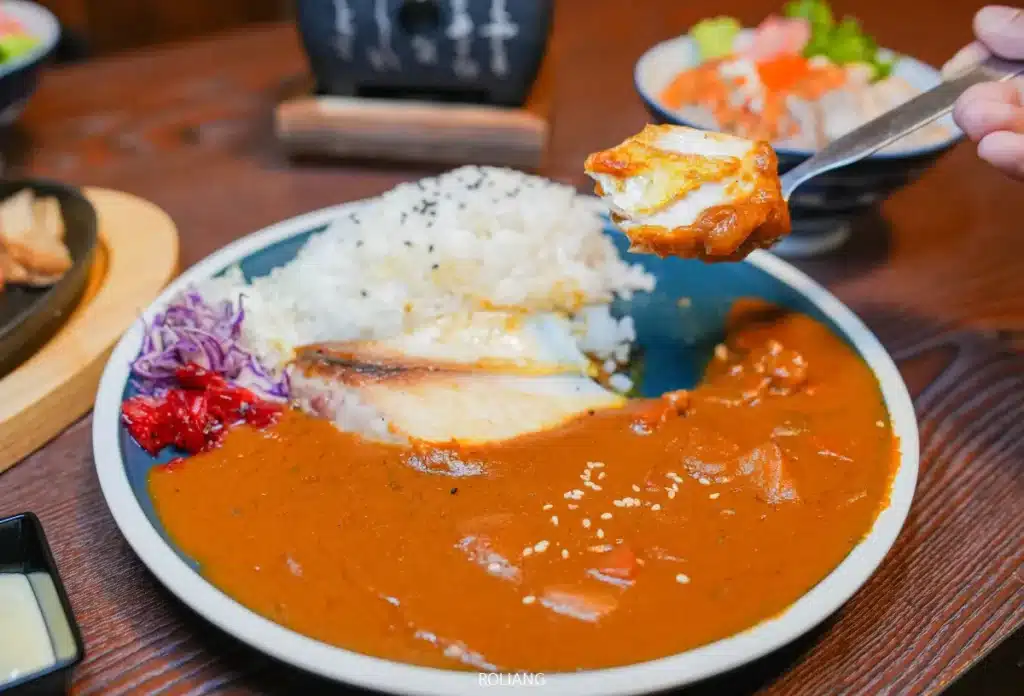 จานแกงกะหรี่ญี่ปุ่นพร้อมข้าวสวย ไก่ชุบเกล็ดขนมปังทอด และผักดอง จากร้าน JAPs ตะกั่วป่า โดยมีช้อนถืออาหารคำหนึ่งอยู่เหนือจาน.