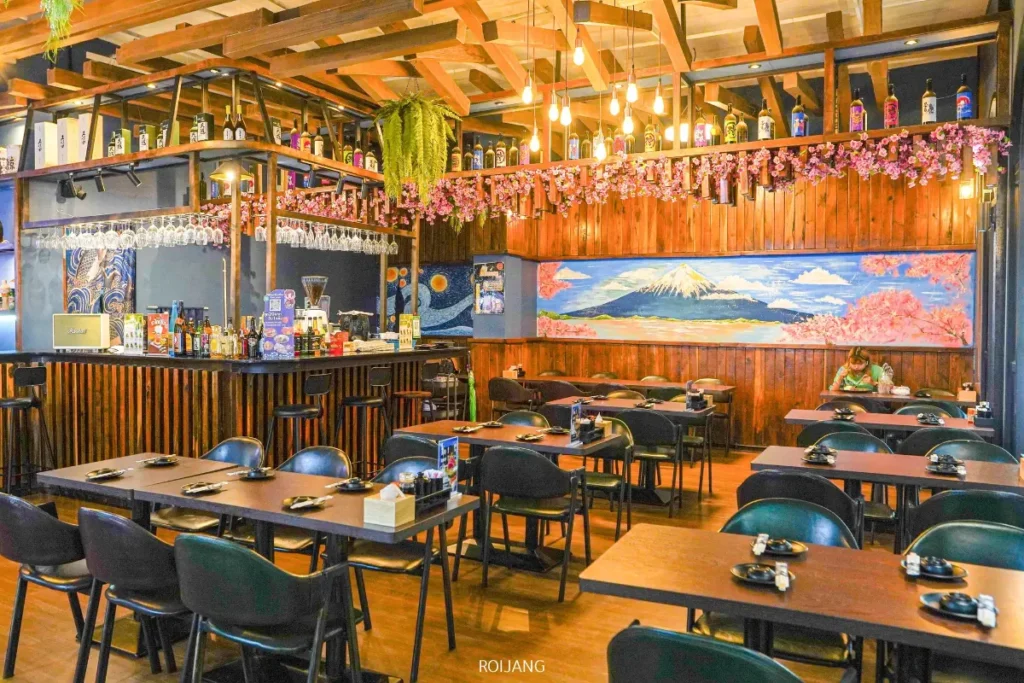 ร้านอาหารสมัยใหม่ที่มีการตกแต่งด้วยไม้ มีแถวของเก้าอี้และโต๊ะสีดำ ภาพจิตรกรรมฝาผนังของภูเขาฟูจิ พืชแขวน และชั้นวางขวดเหนือบริเวณบาร์ที่ JAPs ตะกั่วป่า.