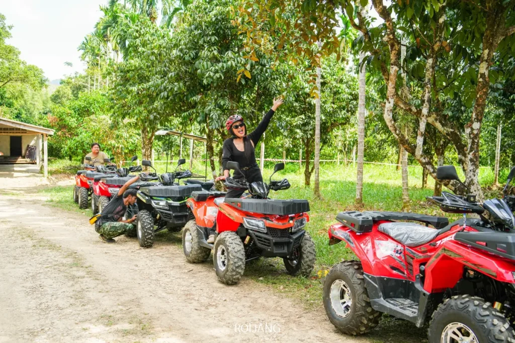 มีผู้คนสี่คนนั่งอยู่บนรถเอทีวีสีแดงที่จอดอยู่บนเส้นทางดินในหุบเขาหมอกเขียวชอุ่ม; มีคนหนึ่งยกแขนขึ้นและยิ้มอยู่.