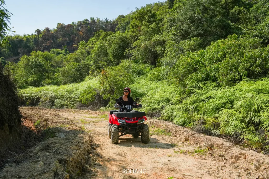 บุคคลหนึ่งขี่รถเอทีวีสีแดงไปตามเส้นทางดินที่คดเคี้ยวผ่านพุ่มไม้เขียวขจีและต้นไม้ในหุบเขาหมอกในวันที่แดดออก.