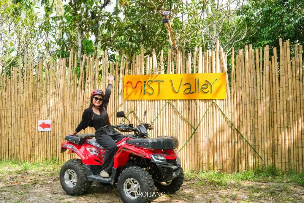 บุคคลที่สวมหมวกนิรภัยนั่งอยู่บนรถเอทีวีสีแดง ยิ้มและยกแขนข้างหนึ่งขึ้นด้านหน้า ถ่ายรูปอย่างภาคภูมิใจใต้ป้าย "หุบเขาหมอก" สีเหลืองซึ่งติดอยู่หน้าแนวรั้วไม้ไผ่.