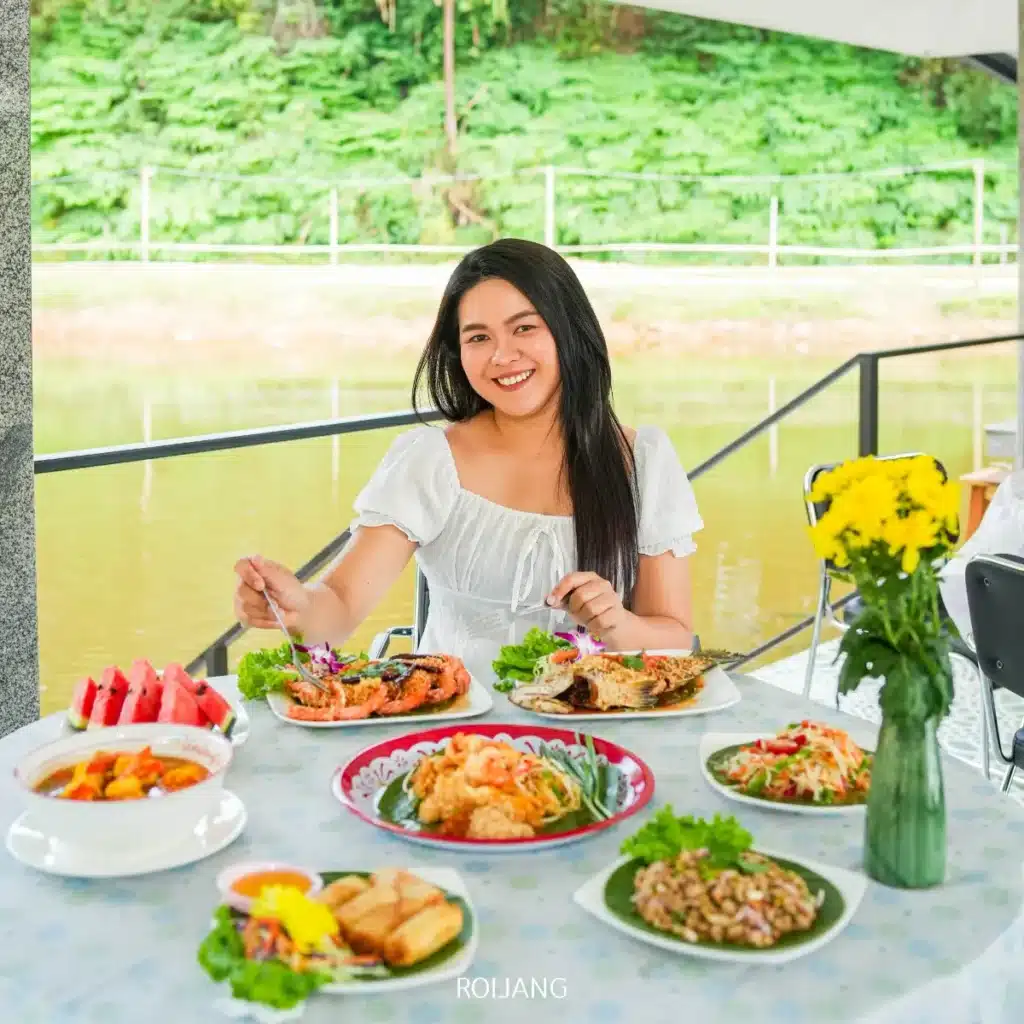 ผู้หญิงในชุดเดรสสีขาว นั่งอยู่ที่โต๊ะกลางแจ้งใกล้หุบเขาหมอก ยิ้มและรับประทานอาหาร พร้อมด้วยอาหารไทยหลากหลายชนิด และแจกันดอกไม้สีเหลืองอยู่ข้างหน้าเธอ.