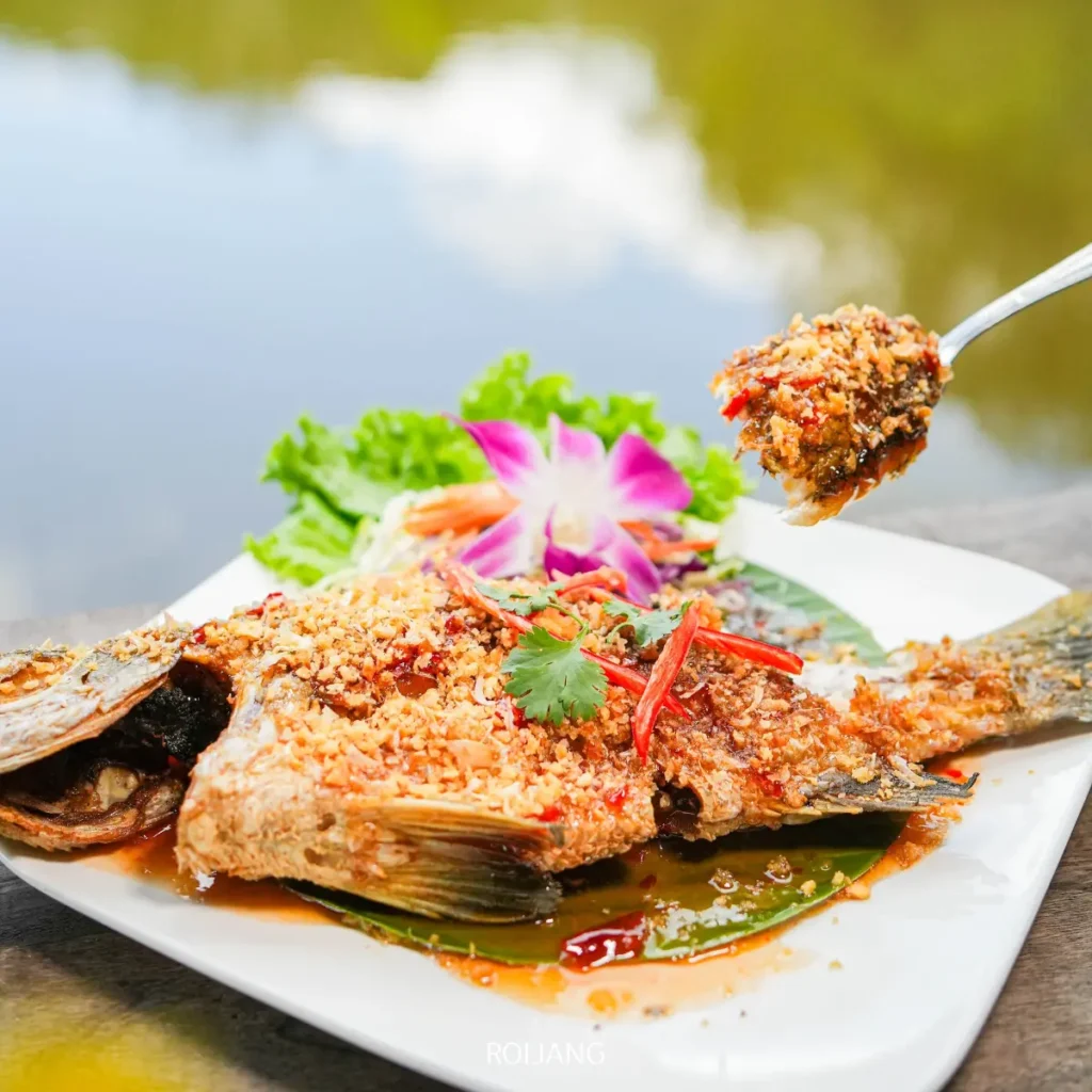 ปลาทอดทั้งตัวราดด้วยซอสกระเทียมและพริกเสิร์ฟบนจานขาว พร้อมผักกาดหอม สมุนไพร ดอกไม้สีม่วงตกแต่งด้านข้าง สร้างสรรค์การนำเสนอที่ได้แรงบันดาลใจจากหุบเขาหมอก.