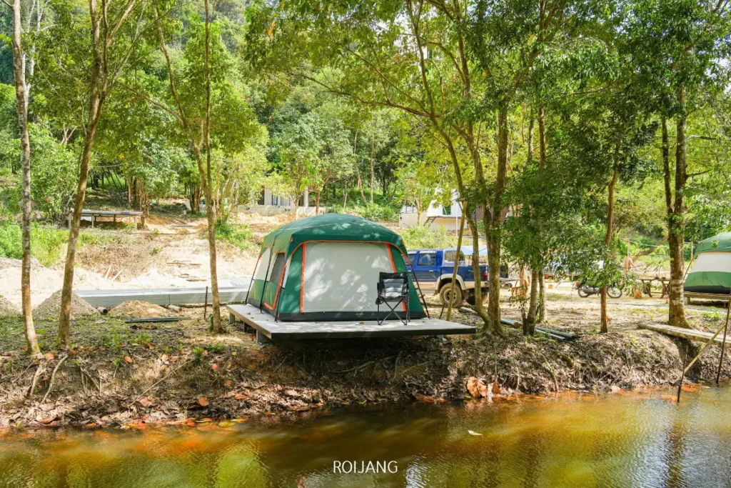 เต็นท์สีเขียวและสีเทาตั้งอยู่บนแท่นไม้ริมแม่น้ำในหุบเขาหมอก ล้อมรอบด้วยต้นไม้ มีเก้าอี้ตัวหนึ่งอยู่ด้านหน้าและยานพาหนะจอดอยู่ด้านหลัง.