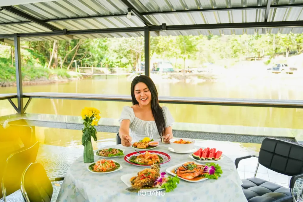 ผู้หญิงคนหนึ่งนั่งอยู่ที่โต๊ะริมแม่น้ำในหุบเขาหมอก ยิ้มและเสิร์ฟอาหารจากจานหลากสีสันหลายจาน รวมถึงผลไม้และอาหารทะเล บนผ้าปูโต๊ะสีขาว.