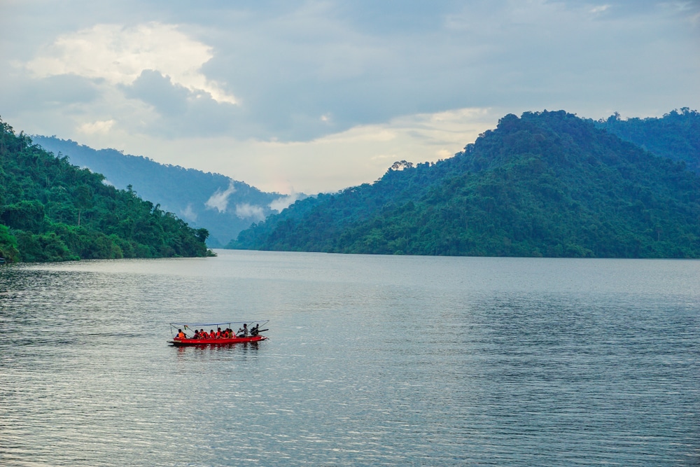 กลุ่มคนในเรือสีแดงกำลังพายข้ามทะเลสาบที่เงียบสงบ รายล้อมไปด้วยภูเขาเขียวขจีภายใต้ท้องฟ้าที่มีเมฆปกคลุม เหมาะสำหรับผู้ที่ เที่ยวใกล้กรุงเทพ ต้องการเที่ยวใกล้กรุงเทพฯ ที่กำลังมองหาการหลบหนีที่เงียบสงบ.