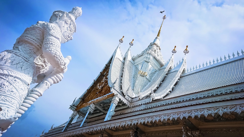 วัดสีขาวประดับประดาอย่างวิจิตรพร้อมลวดลายแกะสลักที่ซับซ้อนและรูปปั้นผู้พิทักษ์อยู่ด้านหน้า ที่เที่ยวใกล้กรุงเทพ ท่ามกลางท้องฟ้าสีฟ้าที่มีนกบินอยู่ด้านบน—สถานที่ที่เหมาะสำหรับผู้ที่ต้องการ เที่ยวใกล้กรุงเทพ และค้นพบสมบัติทางวัฒนธรรมที่ไม่เหมือนใคร.