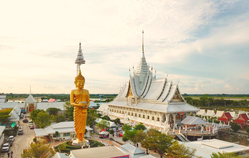 พระพุทธรูปทองคำขนาดใหญ่ตั้งอยู่ข้างพระอุโบสถสีขาวประดับประดาอย่างงดงาม มีหลังคาชั้นลดหลั่น ที่เที่ยวใกล้กรุงเทพ กัน ล้อมรอบด้วยต้นไม้และอาคารต่าง ๆ ภายใต้ท้องฟ้าที่มีเมฆบางส่วน—เหมาะสำหรับผู้ที่ต้องการสถานที่ท่องเที่ยวใกล้กรุงเทพฯ และการพักผ่อนที่สงบเงียบ.