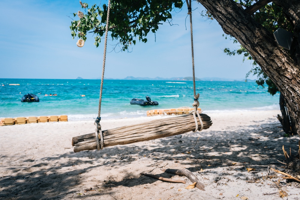 ชิงช้าไม้แขวนอยู่บนต้นไม้บนชายหาดทรายที่มีน้ำทะเลใสสีฟ้าและเรืออยู่ในฉากหลัง ทำให้เป็นสถานที่เหมาะ ที่เที่ยวใกล้กรุงเทพ สำหรับการผ่อนคลายที่ ทะเลใกล้กรุงเทพ.