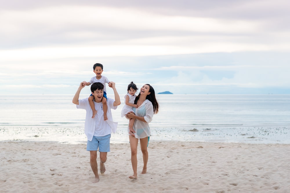 ครอบครัวสี่คนเดินและยิ้มบนชายหาดทรายใกล้ทะเล เที่ยวใกล้กรุงเทพ เพลิดเพลินกับความสงบ ทะเลใกล้กรุงเทพ—สถานที่ท่องเที่ยวธรรมชาติใกล้กรุงเทพที่สมบูรณ์แบบสำหรับทุกคน โดยมีเด็กอยู่บนบ่าของพ่อและแม่กำลังอุ้มเด็กอีกคน.