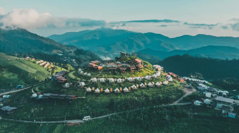ภาพมุมสูงของหมู่บ้านรูปวงกลมที่มีเต็นท์และอาคารตั้งอยู่บนเนินเขาสีเขียว ณ แหล่งท่องเที่ยวม่อนแจ่ม ล้อมรอบด้วยภูเขาภายใต้ท้องฟ้าที่มีเมฆบางส่วน.