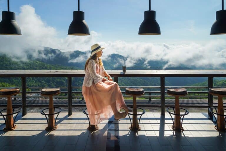 ผู้หญิงคนหนึ่งสวมหมวกและกระโปรงยาว นั่งอยู่คนเดียวที่เคาน์เตอร์พร้อมเครื่องดื่ม มองออกไปยังภูเขาและเมฆผ่านหน้าต่างบานใหญ่ในคาเฟ่สมัยใหม่ เหมาะสำหรับผู้ที่กำลังมองที่พักเพชรบูรณ์ในเมือง หรือโรงแรมเพชรบูรณ์ที่มีสไตล์.