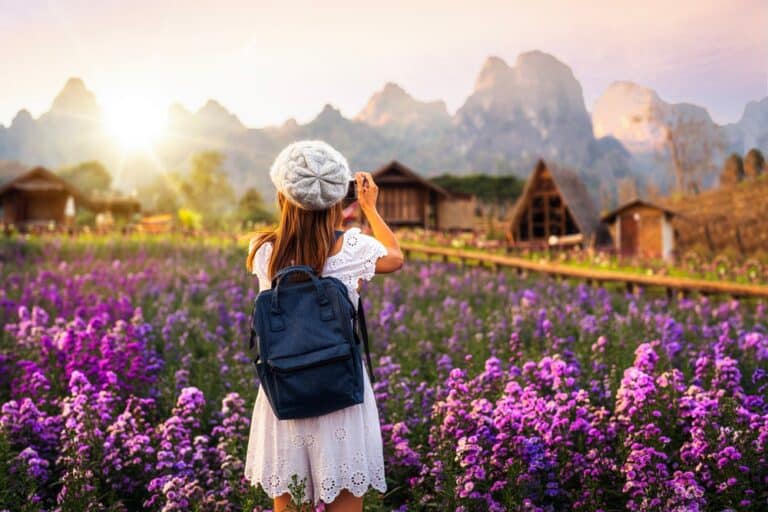 หญิงสาวในชุดเดรสสีขาวสะพายเป้และสวมหมวกเบเรต์กำลังถ่ายภาพในทุ่งดอกไม้สีม่วงยามพระอาทิตย์ตกดิน โดยมีกระท่อมไม้และภูเขาเป็นฉากหลัง—ถ่ายทอดความงดงามอันเงียบสงบของม่อนแจ่ม สถานที่ท่องเที่ยวยอดนิยม.