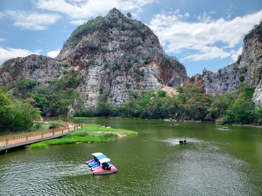ทิวทัศน์อันงดงามของทะเลสาบสีเขียว หนึ่งในสถานที่ท่องเที่ยวธรรมชาติใกล้กรุงเทพฯ ล้อมรอบ เที่ยวใกล้กรุงเทพ ด้วยเนินเขาหิน มีผู้คนพายเรือถีบสีสันสดใสและทางเดินไม้เลียบชายฝั่งใต้ท้องฟ้าที่มีเมฆบางส่วน.
