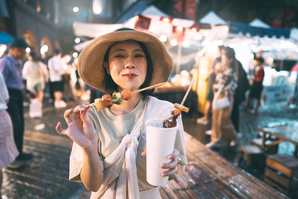 ผู้หญิงคนหนึ่งสวมหมวกถืออาหารเสียบไม้และเครื่องดื่มที่ตลาดกลางคืนกลางแจ้ง ซึ่งเป็นหนึ่งในสถานที่ท่องเที่ยวที่มีชีวิตชีวาของ ที่เที่ยวเชียงใหม่ โดยมีแผงขายของและผู้คนพลุกพล่านในฉากหลังที่เบลอ.