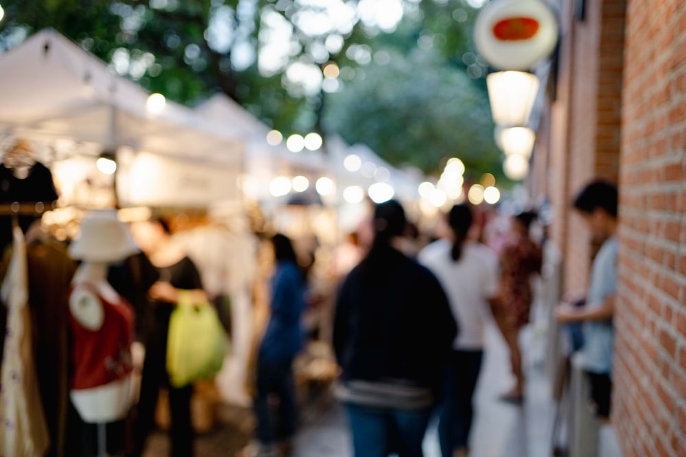 ภาพตลาดกลางแจ้งที่พร่ามัวใน ที่เที่ยวเชียงใหม่ สถานที่ท่องเที่ยว มีผู้คนเดินชมแผงขายของใต้เต็นท์สีขาวและไฟประดับตามถนนที่ล้อมรอบด้วยกำแพงอิฐ.