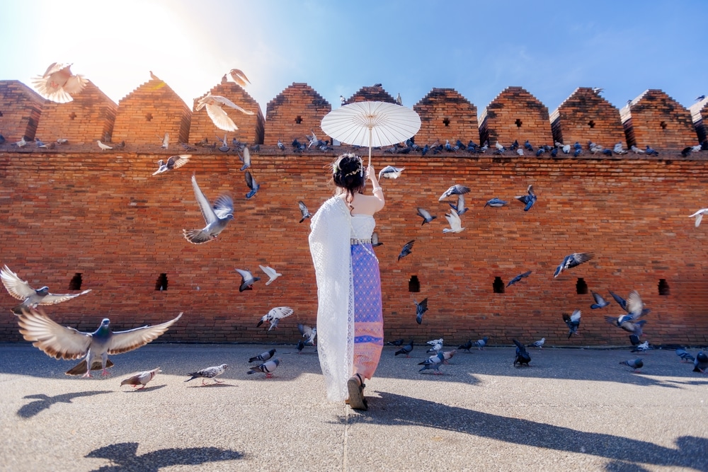 หญิงสาวในชุดพื้นเมืองถือร่มขาวใกล้กำแพงอิฐ ล้อมรอบด้วยนกพิราบที่บินอยู่ใต้ท้องฟ้าแจ่มใส—ถ่ายทอดเสน่ห์ของ ที่เที่ยวเชียงใหม่.