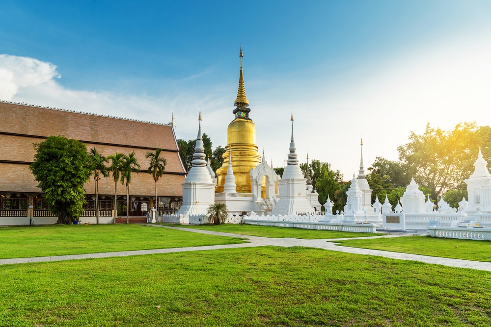 วัดพุทธที่มีพระธาตุสีทองเป็นศูนย์กลาง ล้อมรอบด้วยเจดีย์สีขาว สนามหญ้าสีเขียว และต้นไม้ภายใต้ท้องฟ้าแจ่มใสในยามพระอาทิตย์ตกดิน—สถานที่ท่อง ที่เที่ยวเชียงใหม่ ที่ไม่ควรพลาด.
