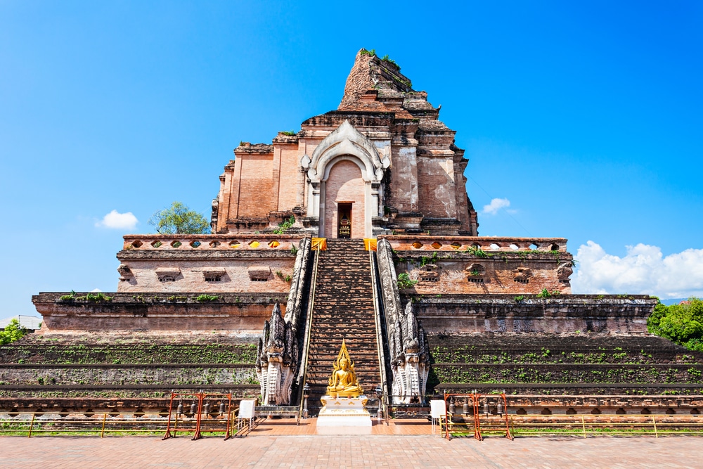 วัดโบราณก่ออิฐที่มีบันไดกลางและพระพุทธรูปทองคำตั้งอยู่ที่ฐาน ตั้งอยู่ใต้ท้องฟ้าสีครามใส—สถานที่ท่อง เที่ยวเชียงใหม่ ที่ต้องไปเยือนสำหรับผู้ที่แสวงหาวัฒนธรรมและประวัติศาสตร์.