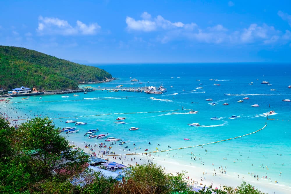 ชายหาดที่แออัดไปด้วยผู้คน น้ำทะเลสีเทอร์ควอยซ์ใสสะอาด เรือ ที่เที่ยวใกล้กรุงเทพ มากมาย และผู้คนกำลังว่ายน้ำ—สถานที่ท่องเที่ยวใกล้กรุงเทพฯ ที่เหมาะอย่างยิ่ง. ภูเขาสีเขียวและท้องฟ้าสีครามสามารถมองเห็นได้ในพื้นหลัง.