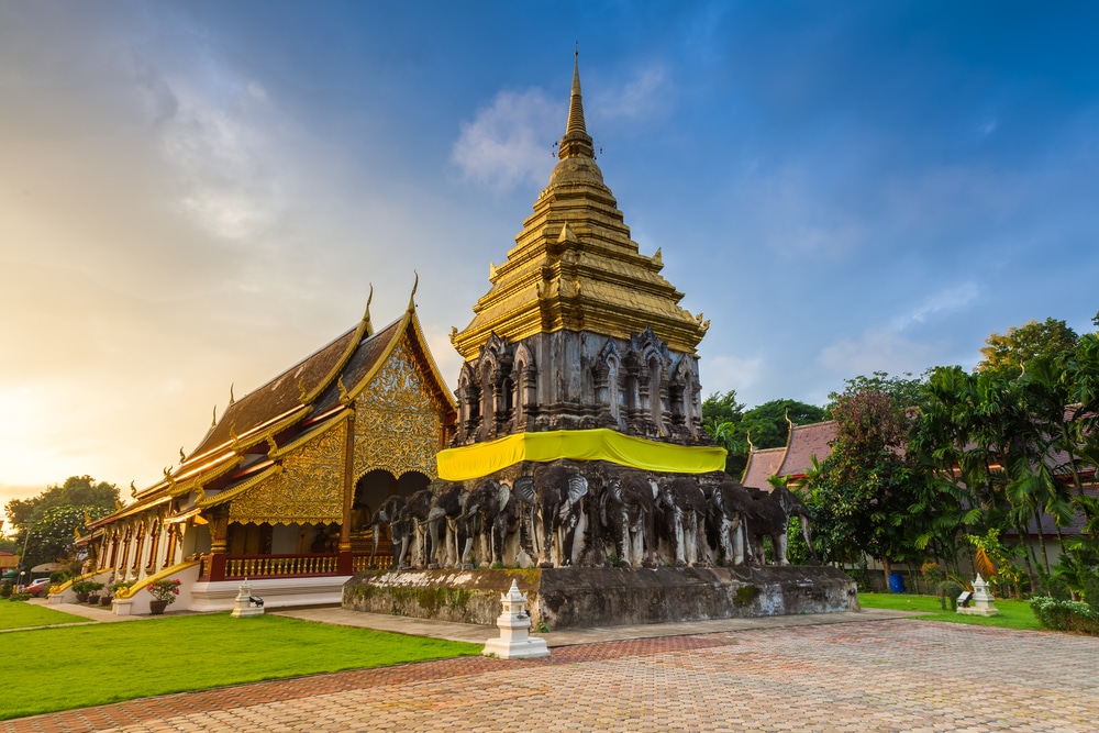 วัดพุทธศาสนา ซึ่งเป็นหนึ่งในสถานที่ท่อง เที่ยวเชียงใหม่ ยอดนิยม ประกอบด้วยเจดีย์ทองคำที่ตั้งอยู่บนฐานซึ่งมีรูปปั้นช้างอยู่ข้างๆ อาคารที่ตกแต่งอย่างวิจิตรภายใต้ท้องฟ้าที่มีเมฆบางส่วน.
