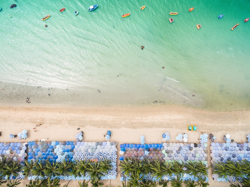 ภาพมุมสูงของชายหาดที่มีร่มเรียงราย เก้าอี้อาบแดด ต้นปาล์ม และ ที่เที่ยวใกล้กรุงเทพ ผู้คนในน้ำสีฟ้าครามพร้อมเรือ—เหมาะสำหรับการพักผ่อนหรือจับคู่ทริปของคุณกับสถานที่ท่องเที่ยวธรรมชาติใกล้กรุงเทพฯ เพื่อการพักผ่อนที่สมบูรณ์แบบ.