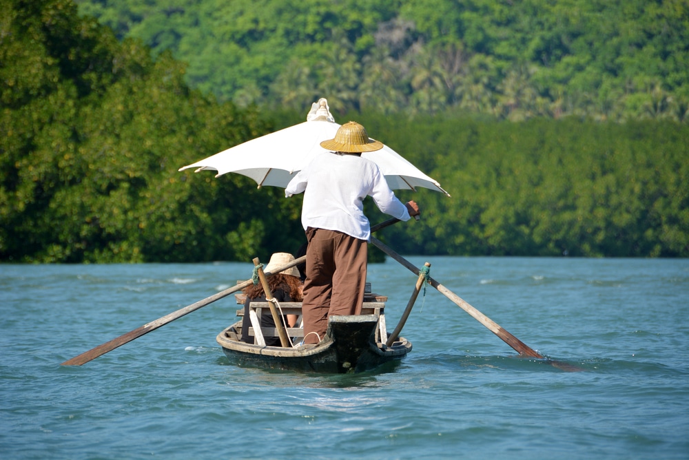บุคคลกำลังพายเรือไม้พร้อมร่มสีขาวบนแม่น้ำ ล้อมรอบด้วยต้นไม้ ที่เที่ยวใกล้กรุงเทพ เขียวชอุ่มและภูเขา—ภาพที่งดงามราวกับฝันสำหรับผู้ที่ต้องการหาสถานที่ท่องเที่ยวธรรมชาติใกล้กรุงเทพฯ เพื่อผ่อนคลายและเติมพลังในความงดงามของธรรมชาติ.