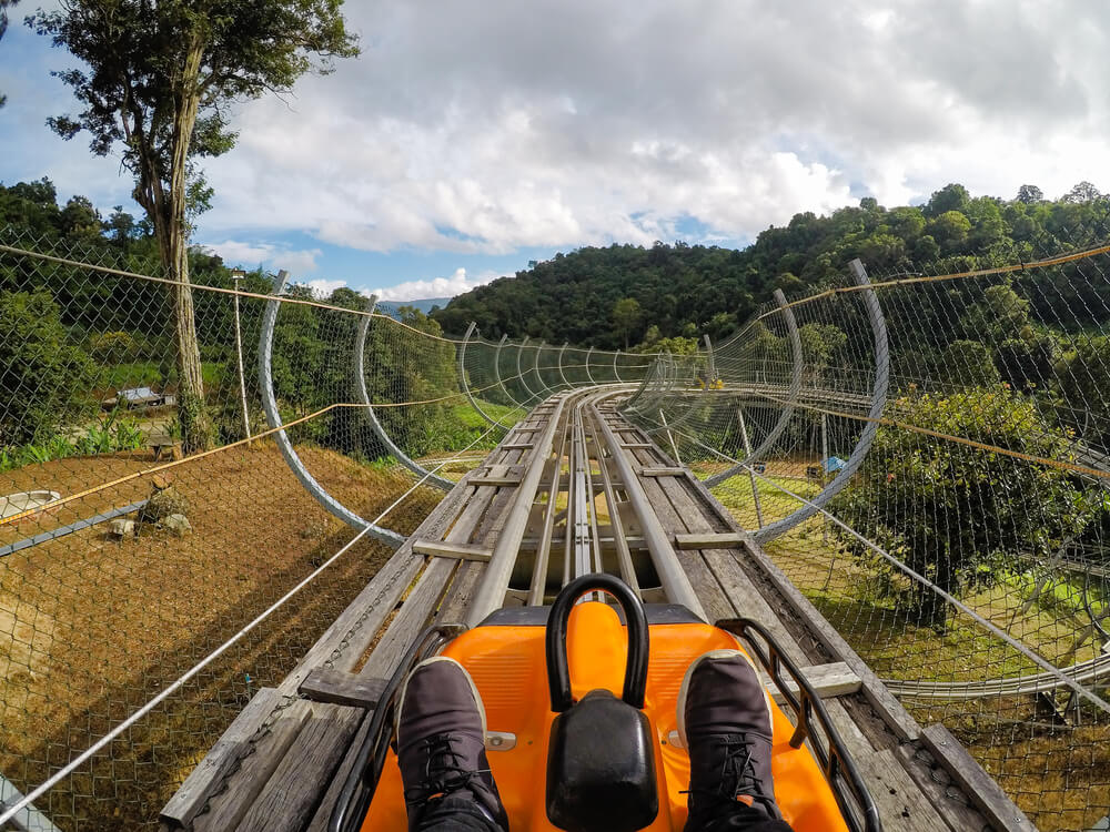 บุคคลหนึ่งนั่งรถไฟเหาะตีลังกาแบบนั่งเดี่ยวสีส้มลงรางไม้และโลหะที่ล้อมรอบด้วยรั้วนิรภัยและเนินเขาสีเขียว—การผจญภัยที่น่าตื่นเต้นใน เชียงใหม่ที่เที่ยว สำหรับผู้แสวงหาความตื่นเต้น.