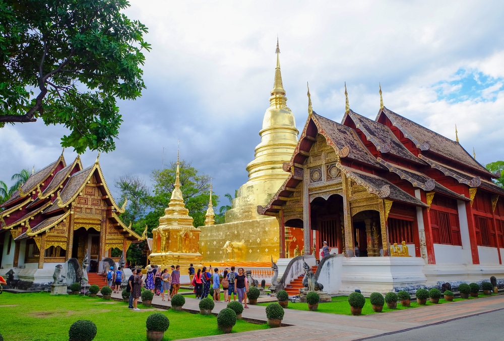 นักท่องเที่ยวมาเยือนวัดพระสิงห์ ซึ่งเป็นสถานที่ท่องเที่ยวที่ต้องไปเยือนใน ที่เที่ยวเชียงใหม่ มีเจดีย์ทองคำและอาคารไม้ที่ตกแต่งอย่างวิจิตรภายใต้ท้องฟ้าที่มีเมฆปกคลุม.