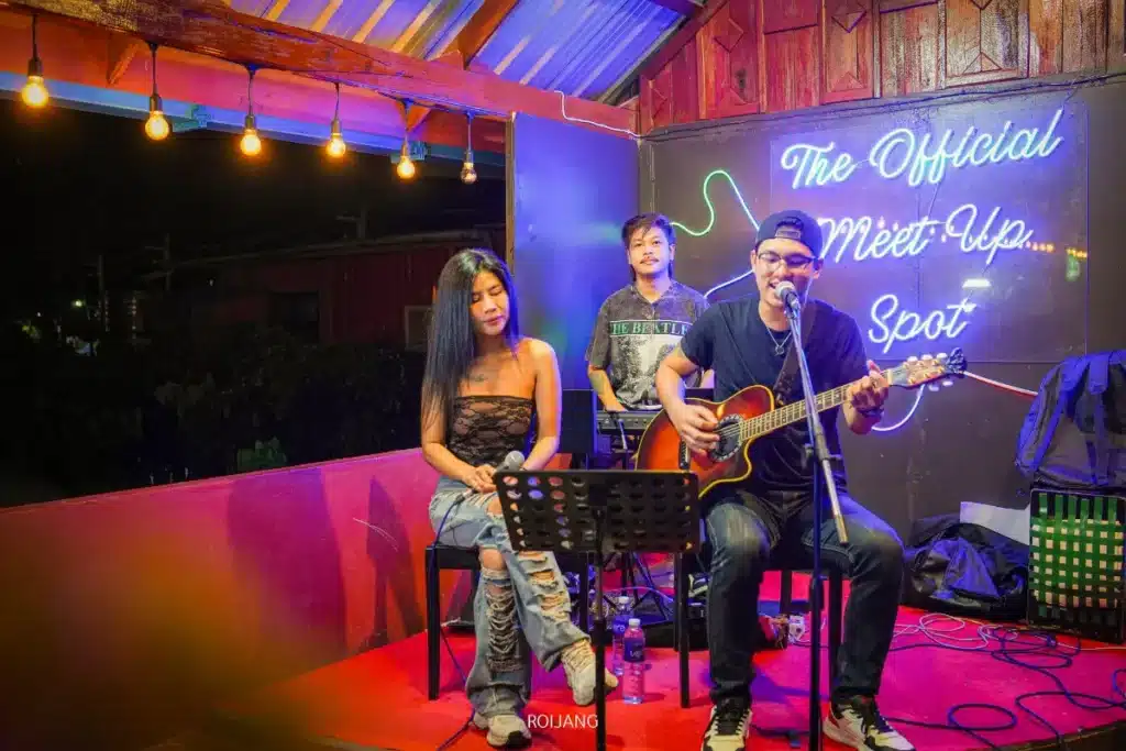 นักดนตรีสามคนกำลังแสดงบนเวทีเล็ก ๆ ในร่มที่ The Rooftop; ผู้หญิงคนหนึ่งนั่งอยู่กับไมโครโฟน ผู้ชายคนหนึ่งเล่นกีตาร์อะคูสติกและร้องเพลง และผู้ชายอีกคนหนึ่งเล่นคีย์บอร์ด. ป้ายนีออนส่องสว่างอยู่เบื้องหลังพวกเขา.