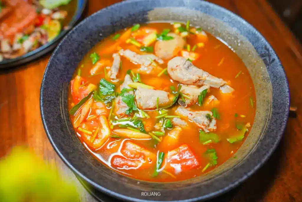 ชามซุปที่มีชิ้นเนื้อ มะเขือเทศ สมุนไพร และผักต่าง ๆ ในน้ำซุปใสสีแดง เสิร์ฟบนโต๊ะไม้ที่ The Rooftop.