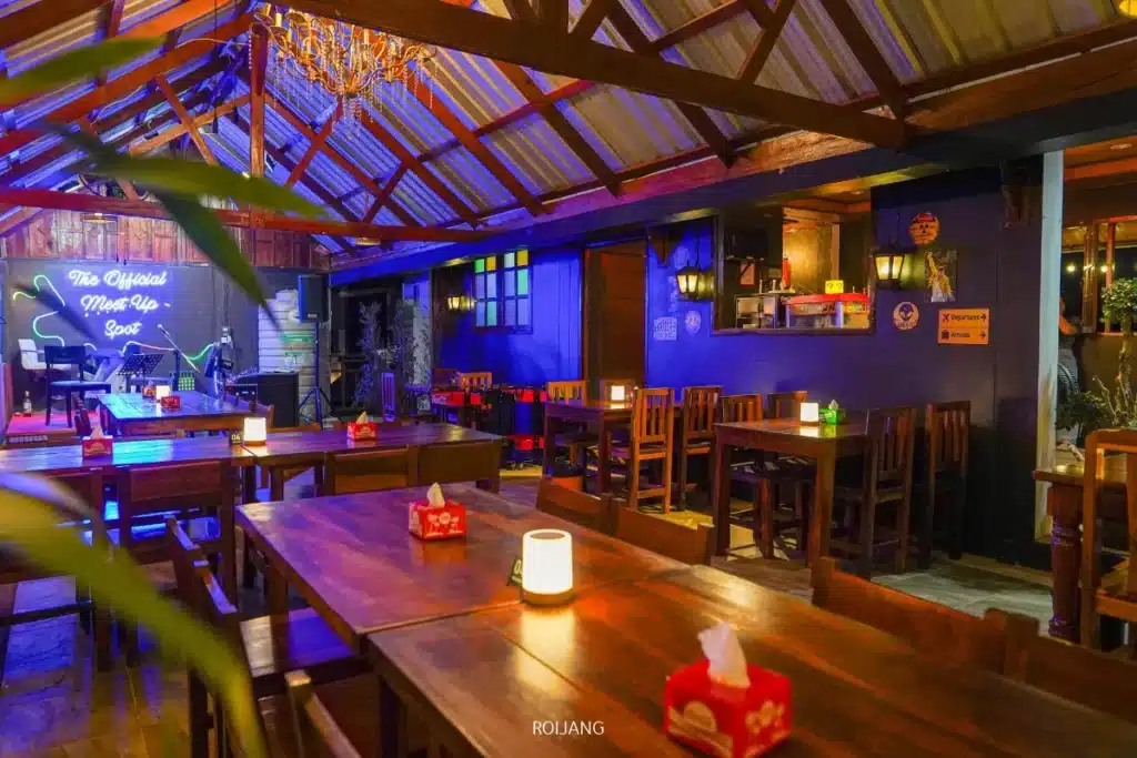 ร้านอาหารบรรยากาศอบอุ่นและแสงสลัวชื่อ เดอะ รูฟท็อป มีโต๊ะและเก้าอี้ไม้ เทียนขนาดเล็กวางอยู่บนโต๊ะทุกโต๊ะ และป้ายไฟนีออนที่ส่องแสงอยู่บนผนังด้านหลังใกล้กับเวทีเล็กๆ.