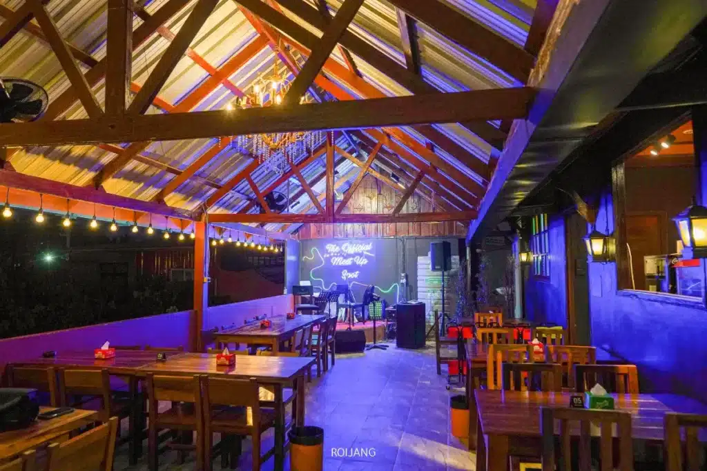 บาร์บนดาดฟ้าประกอบด้วยโต๊ะและเก้าอี้ไม้, ไฟประดับ, และเวทีเล็กสำหรับดนตรีสด ทั้งหมดตั้งอยู่ใต้หลังคาเอียงเพื่อสร้างบรรยากาศในยามค่ำคืนที่อบอุ่น.