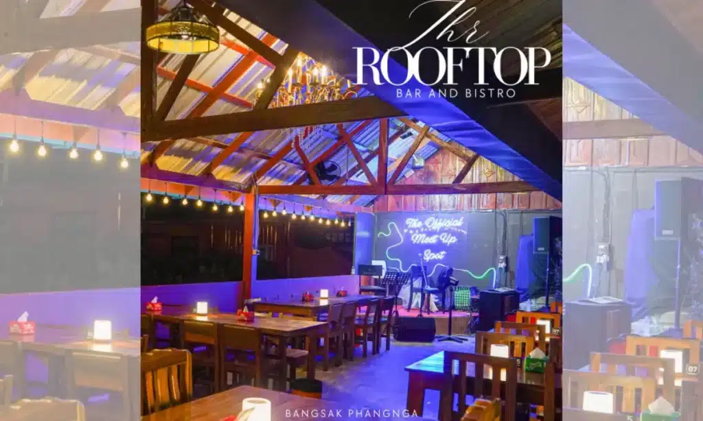 ภายในของ The Rooftop ตกแต่งด้วยโต๊ะและเก้าอี้ไม้ เวทีพร้อมเครื่องดนตรี ป้ายไฟนีออน และแสงไฟอบอุ่น สร้างบรรยากาศที่เชิญชวนซึ่งเป็นเอกลักษณ์ของ The Rooftop Bar and Bistro.