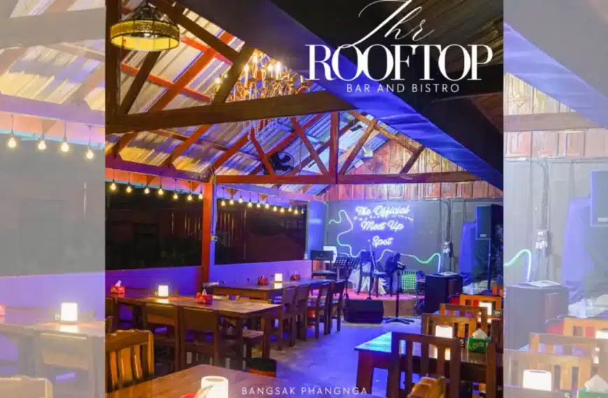 ภายในของ The Rooftop ตกแต่งด้วยโต๊ะและเก้าอี้ไม้ เวทีพร้อมเครื่องดนตรี ป้ายไฟนีออน และแสงไฟอบอุ่น สร้างบรรยากาศที่เชิญชวนซึ่งเป็นเอกลักษณ์ของ The Rooftop Bar and Bistro.