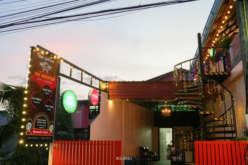 ทางเข้าบาร์และบิสโทรบนดาดฟ้าโดดเด่นด้วยบันไดเวียน ป้ายไฟประดับ ไฟประดับสาย และรั้วสีแดงที่เรืองแสงยามพลบค่ำ.