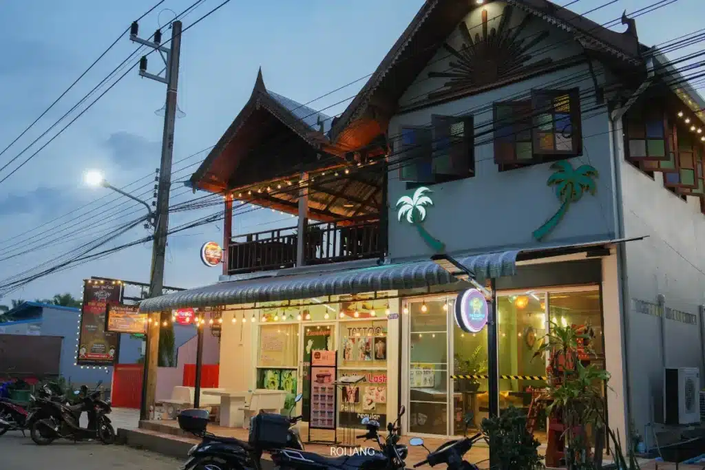 อาคารสองชั้นพร้อมหลังคาตกแต่งที่ The Rooftop มีไฟประดับ สายไฟประดับ โต๊ะนั่งกลางแจ้ง จอดรถจักรยานยนต์ด้านหน้า และมีป้ายไฟสว่างในยามพลบค่ำ.