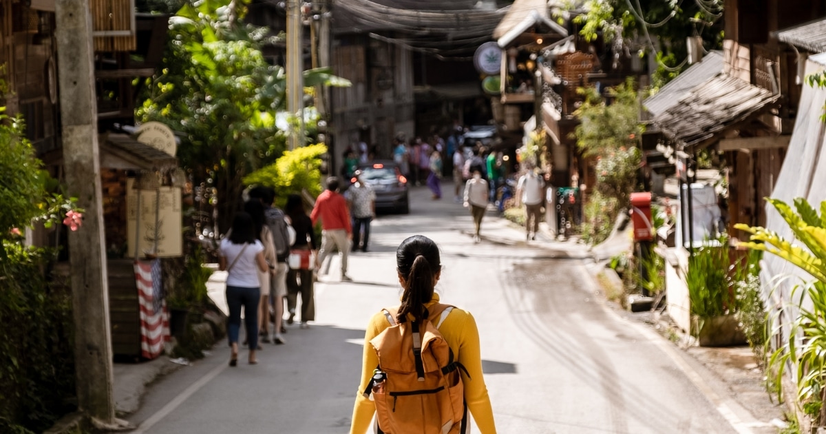 คนที่มีกระเป๋าเป้สีเหลืองเดินไปตามถนนที่พลุกพล่านซึ่งเรียงรายไปด้วยผู้คน อาคาร และต้นไม้ในวันที่มีแดดจัด สำรวจสถานที่ท่องเที่ยวเชียงใหม่.